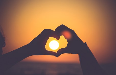 Two hands form a silhouette heart in front of a setting sun